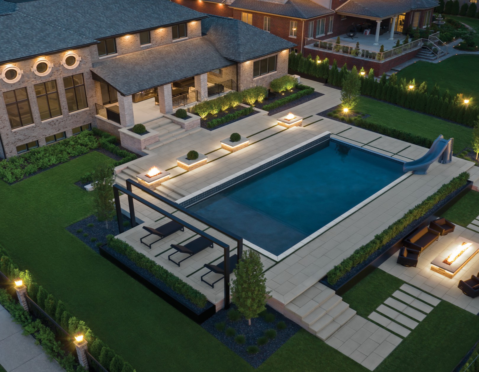 Aerial shot of a landscaped backyard featuring a rectangular pool, large concrete paver patio, lounge area, fire features, and structured plant beds.