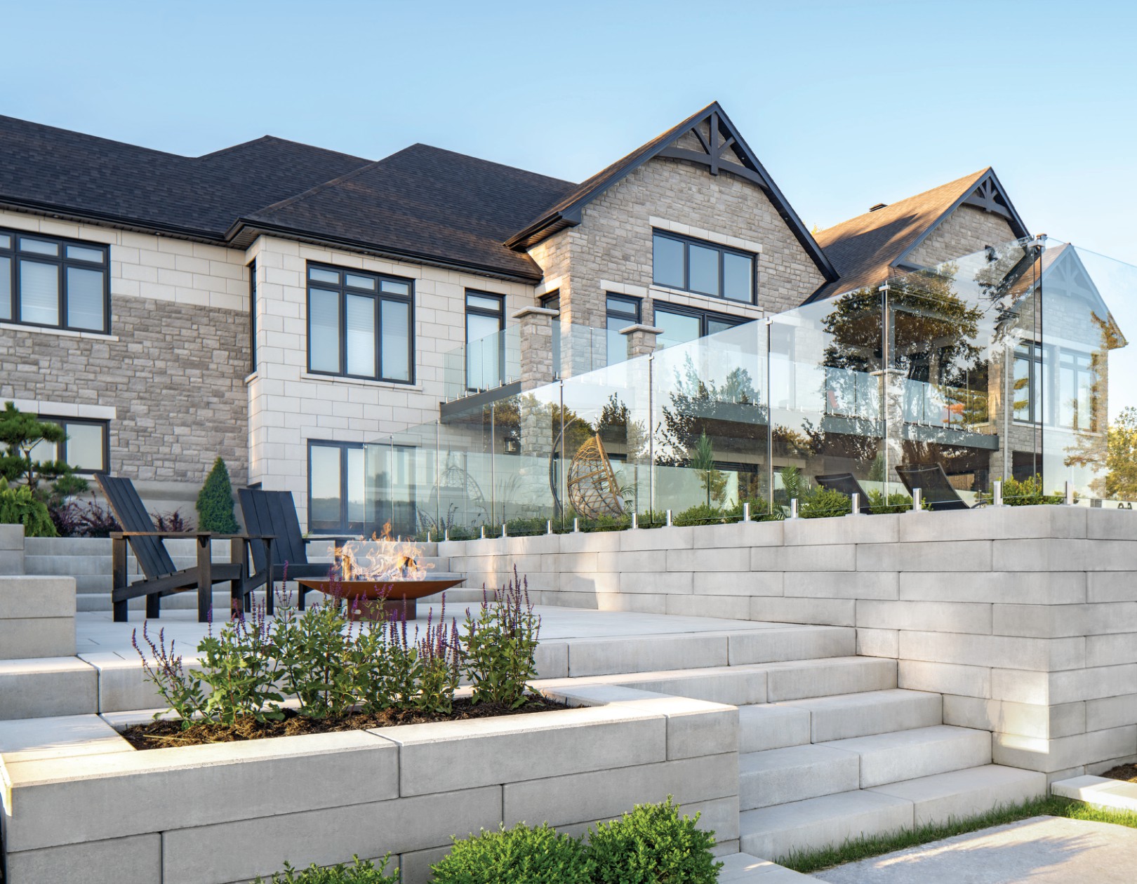 Multi-level outdoor living space featuring stone retaining walls, steps, glass railing, lounge chairs, and a fire pit in front of a large home.