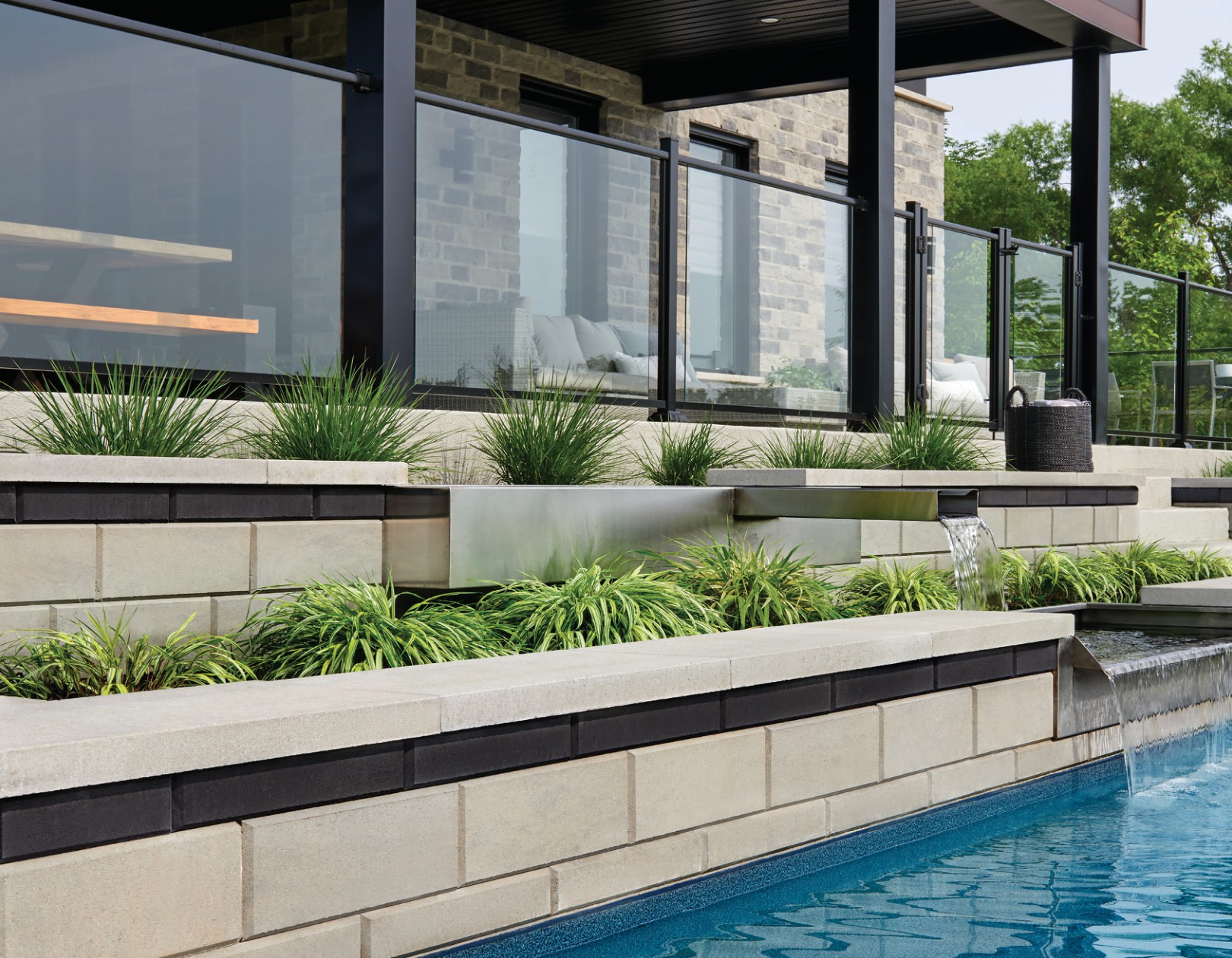Layered stone planter beds with ornamental grasses and a stainless steel spillway water feature flowing into a swimming pool.