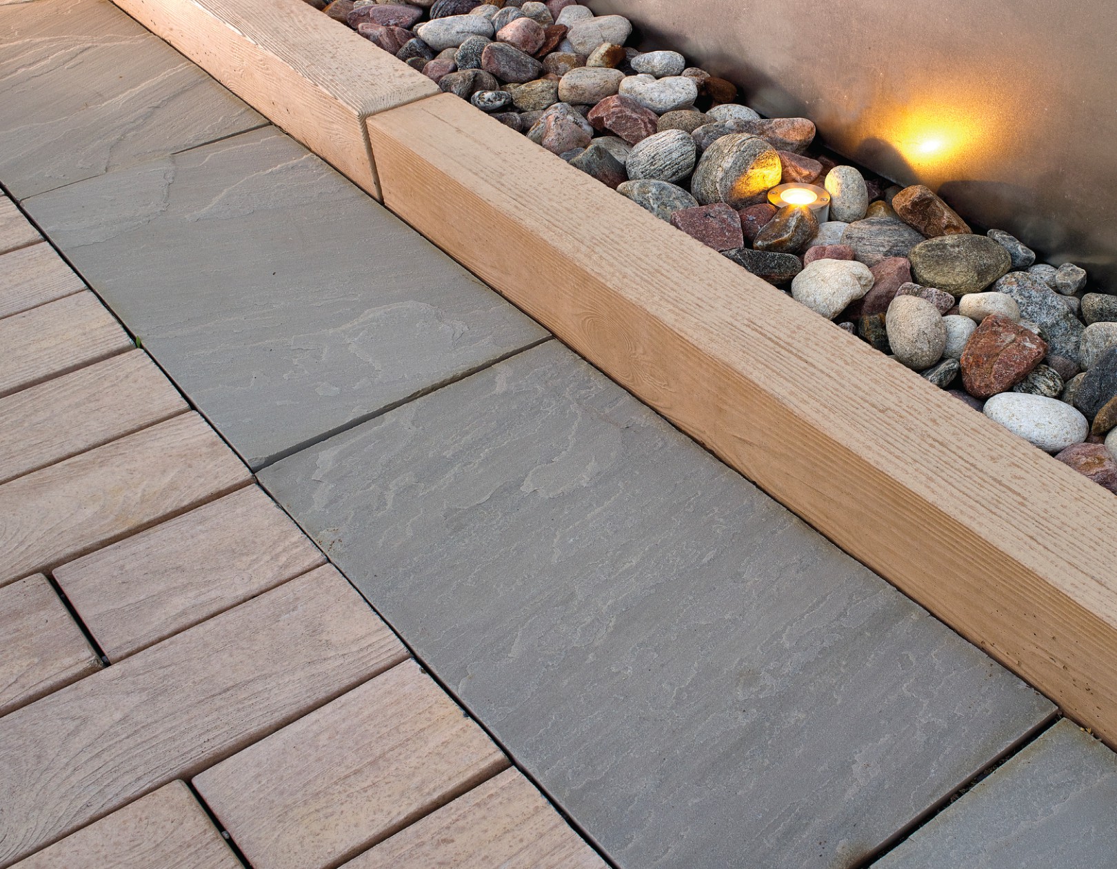Close-up of natural stone pavers bordered by wooden edging and a bed of smooth decorative river rocks with integrated landscape lighting.