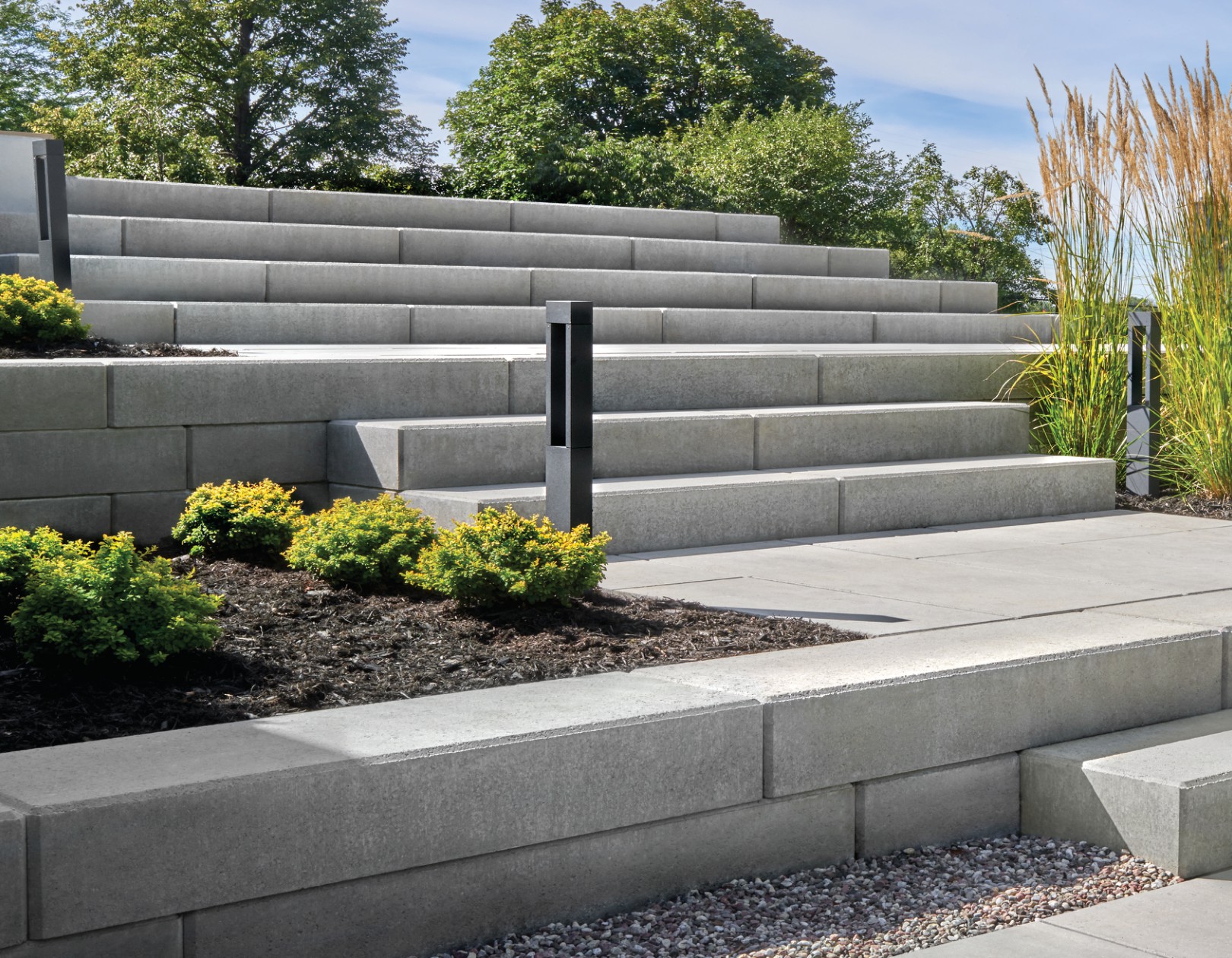 Large outdoor staircase built with smooth concrete blocks, surrounded by retaining walls, shrubs, and tall ornamental grasses.